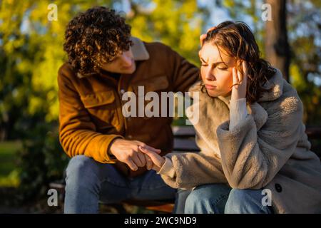 Young couple is sitting in park on sunny day. Woman is sad and man in consoling her. Stock Photo