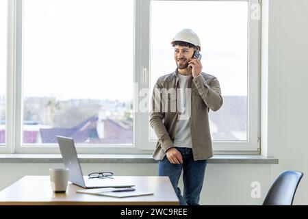 Contractor talking on cell phone while checking laptop and taking notes of what he talks about in the work office. Work organization concept, telecomm Stock Photo