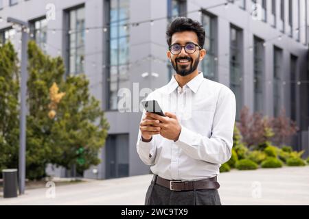 Confident young businessman with a beard, smiling while using smartphone outdoors, office building backdrop. Stock Photo