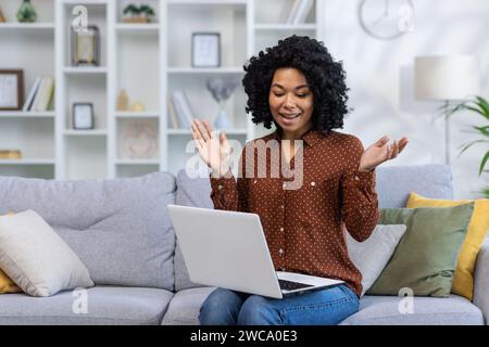 African American young woman sitting on sofa at home and talking smiling and emotional on video call online. Stock Photo