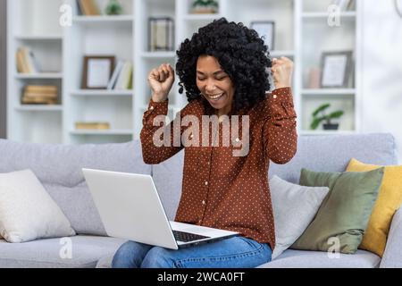 Happy young African American woman is happy with success and victory, raising her hands up and celebrating. Sitting at home on the sofa and looking at the laptop screen. Stock Photo