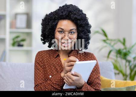 Close-up portrait of a young African-American female teacher teaching remotely from home, sitting on a couch, holding a notebook and a pen, smiling and looking at the camera. Stock Photo