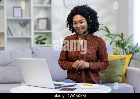 African American young woman wearing a headset sitting on the couch at home and talking on a video call on a laptop. Stock Photo