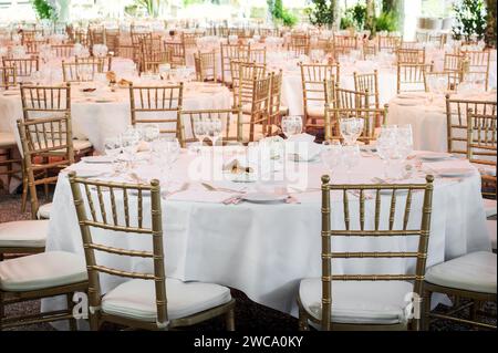 Round tables with white tablecloths and served glassware with golden chairs placed outdoors before event celebration Stock Photo