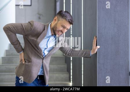 Adult man in smart casual attire experiencing sudden back pain, grimacing in discomfort, leaning on a wall indoors. Stock Photo
