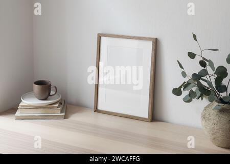 Breakfast still life. Cup of coffee, books. Empty wooden picture frame mockup on desk, table. Textured vase with silver eucalyptus branches. Elegant Stock Photo