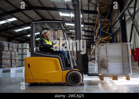 Side view of forklift in warehouse with male driver. Warehouse worker preparing products for shipmennt, delivery, checking stock in warehouse. Stock Photo