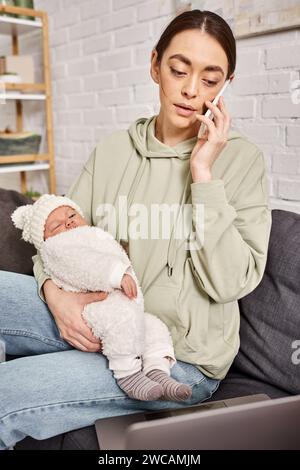 attractive woman in casual attire looking at laptop and holding her baby while talking by phone Stock Photo