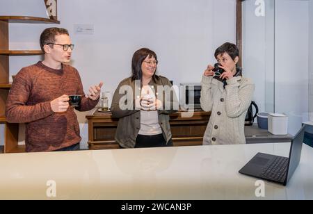 Three co-workers a man and two women having a conversation while they consult a laptop and have coffee on a break from work in office. Workaholics con Stock Photo