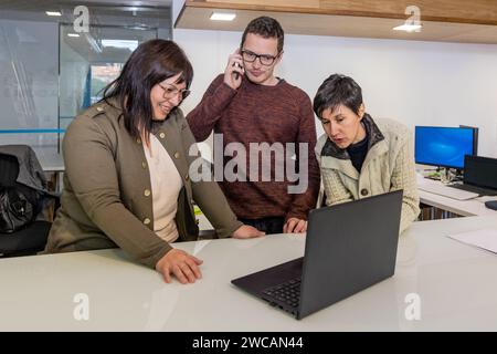 Three co-workers in an office, a man and two women doing a laptop conference at the same time as the man speaks on mobile phone Stock Photo