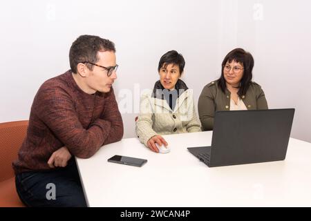 Three co-workers from an office having a conversation in a meeting room. They are sitting at a table on which there is a laptop and a smartphone. Stock Photo