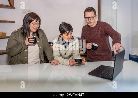 Three co-workers a man and two women having a conversation while they consult a laptop and have coffee on a break from work in the office. Workaholics Stock Photo