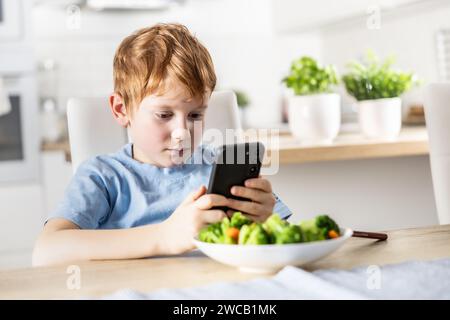 View of smart cute boy using smartphone before lunch in kitchen at home. Stock Photo