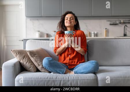 Contented woman savoring warm beverage, seated comfortably on grey sofa Stock Photo