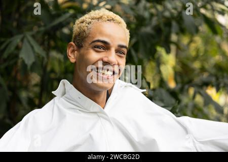 Candid shot of a happy young man with curly blonde hair, laughing in a white shirt, set against a backdrop of dense garden foliage. Stock Photo