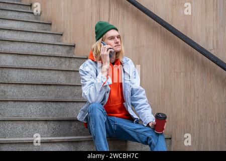Hipster guy with coffee cup in hand sits on steps talks on smartphone in city street.  Stock Photo
