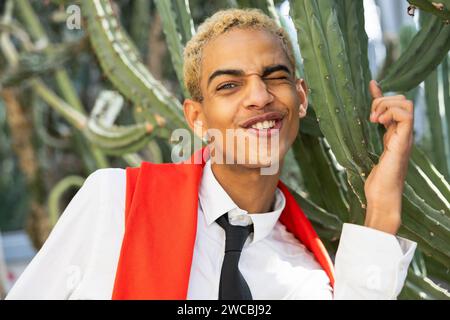 A cheerful young man winks and sticks out his tongue, playfully posing by a large cactus at a botanical garden. Stock Photo