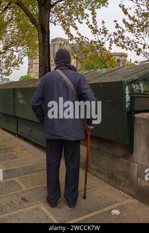 Paris, France, 2019. An elderly man with a cane standing in front of the closed box of a bouquiniste (bookseller) is looking at Notre-Dame de Paris Stock Photo