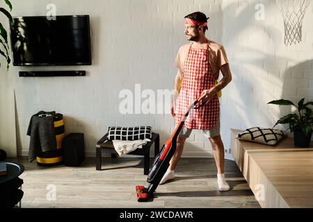 Young serious man in apron standing by wooden cabinets and using electric vacuum cleaner while looking through window Stock Photo