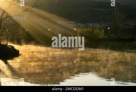 horizontal photograph of a ray of sunlight illuminating a river in the early hours of the day, you can see the mist coming out of the water and the co Stock Photo