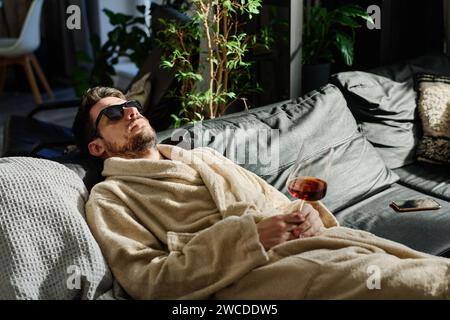 Young man in bathrobe and sunglasses daydreaming with glass of red wine in hand while relaxing on couch with pillows Stock Photo
