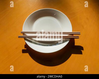 An empty white plate stands on a table. There are two pairs of chopsticks on the plate. The plate and chopsticks cast an interesting shadow on the tab Stock Photo