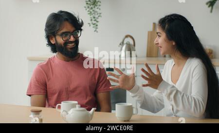 Happy multiracial couple drink coffee tea in home kitchen friendly chatter Arabian man guy listen funny story carefree conversation Indian woman girl Stock Photo