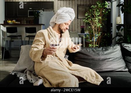 Young relaxed man in bathrobe holding glass of red wine and texting in smartphone or watching online video while sitting on couch Stock Photo