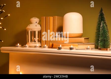 Mantelpiece with books, burning candles and mini Christmas tree near green wall at evening, closeup Stock Photo