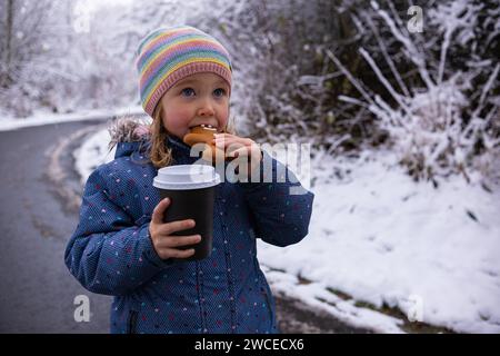 Little cute girl with a cup of tea and a gingerbread man on a snowy winter day. Girl in warm jacket drinking tea and eating gingerbread in the form of Stock Photo