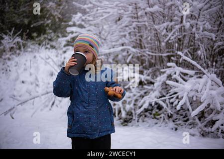 Little cute girl with a cup of tea and a gingerbread man on a snowy winter day. Girl in warm jacket drinking tea and eating gingerbread in the form of Stock Photo