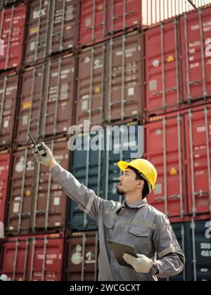 Asian worker wearing safety helmet and using walkie talkie in front of container cargo freight ship for Logistic Import Export background. Stock Photo