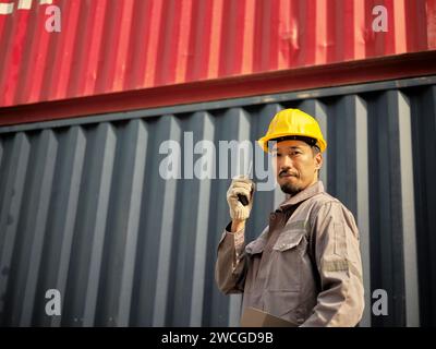 Asian worker wearing safety helmet and using walkie talkie in front of container cargo freight ship for Logistic Import Export background. Stock Photo