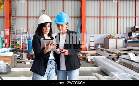Two women wearing hard hats are standing in a warehouse, inspecting the inventory or supervising the operations. Stock Photo