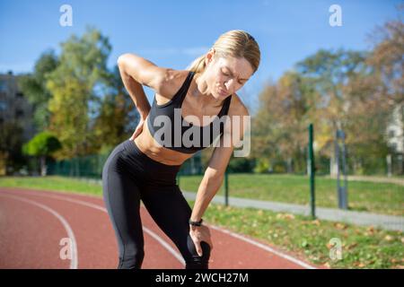 A fit woman in sports attire pauses with hands on knees, exhausted after a strenuous run on a sunny track. Stock Photo