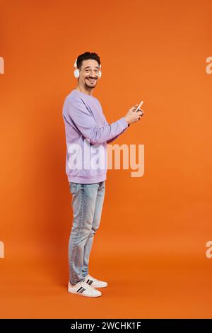 joyful man in wireless headphones listening music and using smartphone on orange background Stock Photo