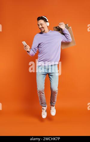 excited man in headphones levitating with shopping bags and holding smartphone on orange background Stock Photo