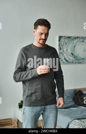 bearded man grimacing while suffering from migraine and holding glass of water at home, unwell Stock Photo