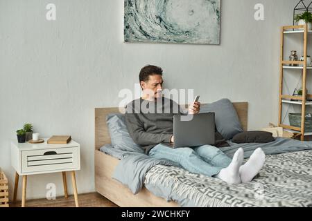 bearded man using laptop and holding mobile phone while working remotely from home, freelance Stock Photo