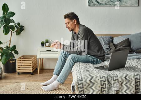 bearded man using mobile phone while working remotely from home near laptop on bed, freelance Stock Photo