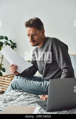 focused and bearded man holding smartphone and document while using laptop, remote work Stock Photo