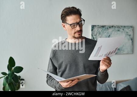 focused and bearded man in eyeglasses and grey sweater analyzing graphs while working from home Stock Photo