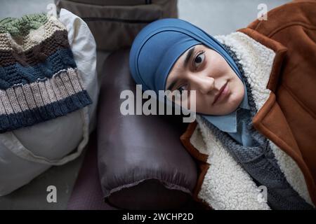 Close up portrait of young Middle Eastern woman wearing headscarf lying on floor in refugee shelter and looking at camera with striking dark eyes, copy space Stock Photo