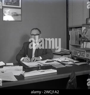 1970s, historical, a business executive in the motor trade, at his desk with paperwork and pen in hand, an auto dealer, possibly a company accountant, as on the desk is a brochure about national insurance contributions. On the right a phone system of the era, with a phone hanging on a stand with dial switch below. Stock Photo