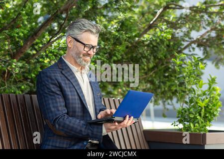 Elegant senior businessman with glasses using a digital tablet while sitting on a bench surrounded by greenery. Stock Photo
