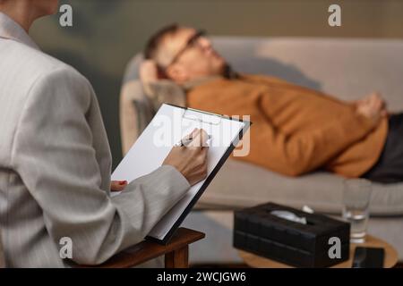 Close up of therapist holding blank clipboard and taking notes with client lying on couch in background, copy space Stock Photo