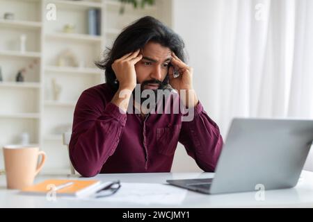 Concerned young indian man looking at laptop screen and holding his temples Stock Photo