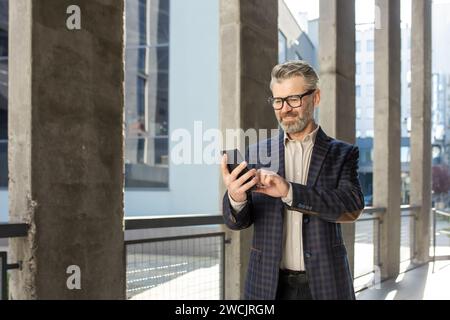 Smiling senior businessman in a stylish suit engaged with his smartphone outside a contemporary office setting. Stock Photo