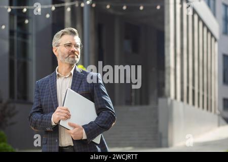 Mature executive in trendy suit holding a laptop while walking outside a modern office building, exuding confidence and professionalism. Stock Photo