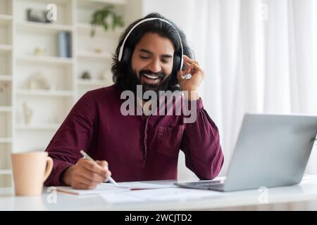 Distance Learning. Smiling Indian Man In Headphones Using Laptop And Taking Notes Stock Photo